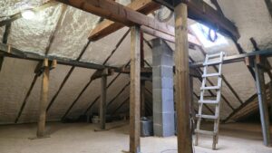Izolacja poddasza z użyciem pianki PUR, ukazująca drewniane belki i konstrukcję dachu. Ocieplanie poddasza przyczynia się do zwiększenia efektywności energetycznej domu. W tle widoczna drabina oraz elementy przygotowujące do dalszych prac izolacyjnych.