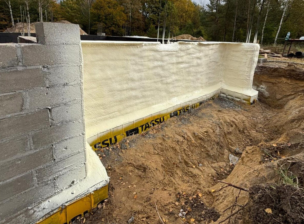 Izolacja fundamentów budynku wykonana przy użyciu piany PUR, ukazująca proces ocieplania, który poprawia efektywność energetyczną domu. Widoczne warstwy materiału izolacyjnego na etapie budowy.