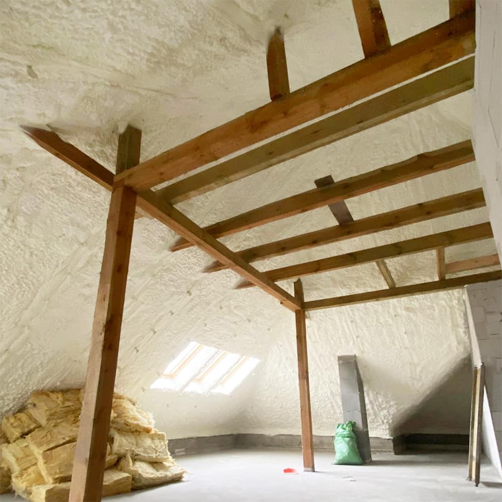 Izolacja poddasza wykonana pianą PUR, widok na drewniane krokwie i ocieplanie poddasza. W tle materiały do izolacji fundamentów, co zwiększa efektywność energetyczną domu.